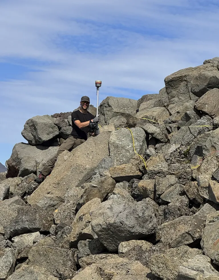 Daníel Ben-Yehoshua skriðusérfræðingur stendur í stórgrýttri urð í Strandartindi og heldur á mælitæki. Mælibúnaður og kapall liggja á milli steina.